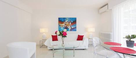 Una sala de estar moderna con una decoración blanca, una mesa de centro de vidrio, un sofá con cojines rojos y un cuadro en la pared.