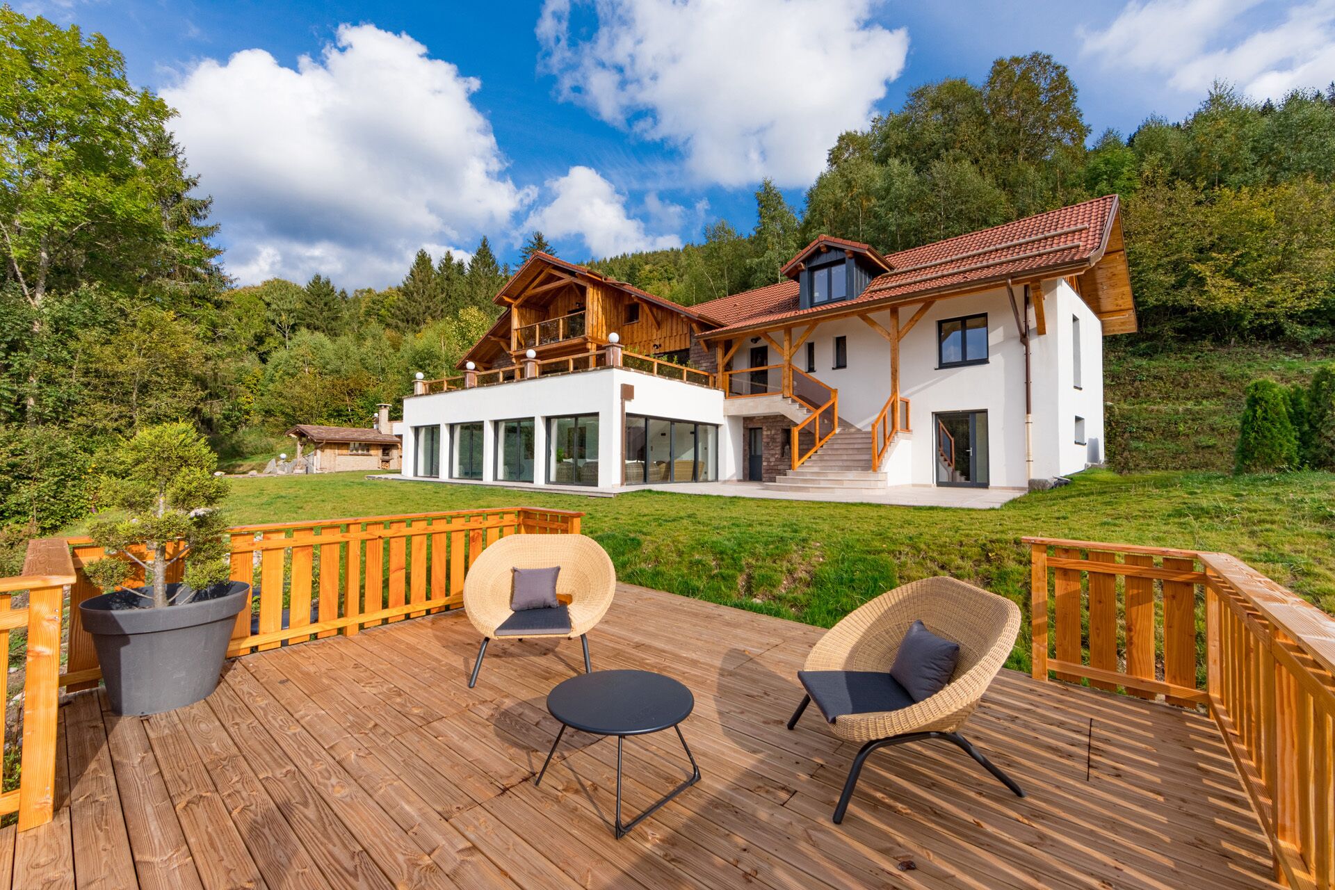 Een modern, twee verdiepingen tellend huis met een houten terras, buitenmeubels en een groot raam.