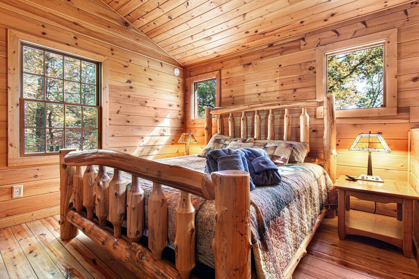 A cabin with a wooden interior, a bed with a headboard, two windows, and a nightstand with a lamp.