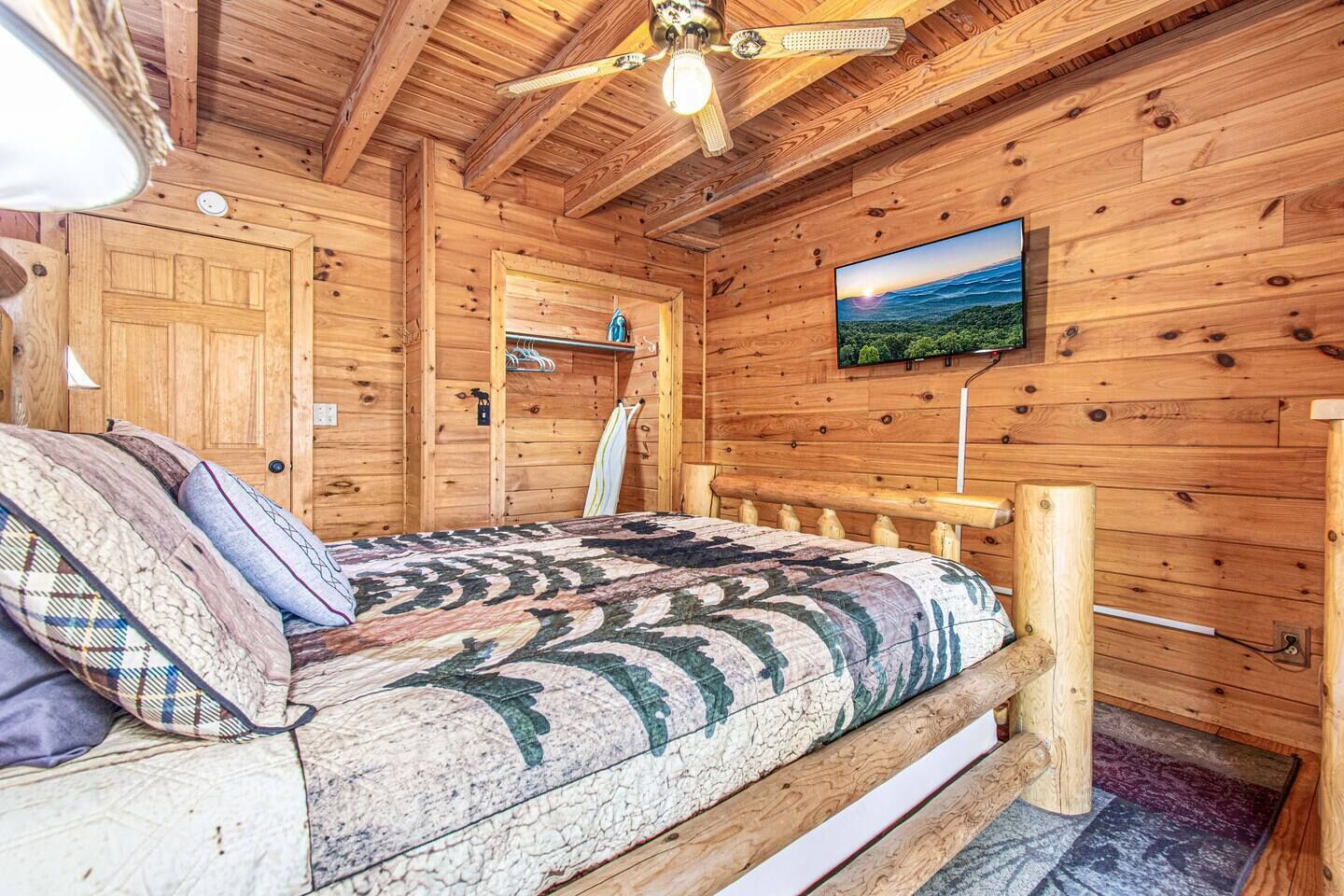 Une chambre dans un chalet en bois, équipée d’un lit, d’un ventilateur de plafond, d’un téléviseur fixé au mur et d’une porte avec un porte-manteau.