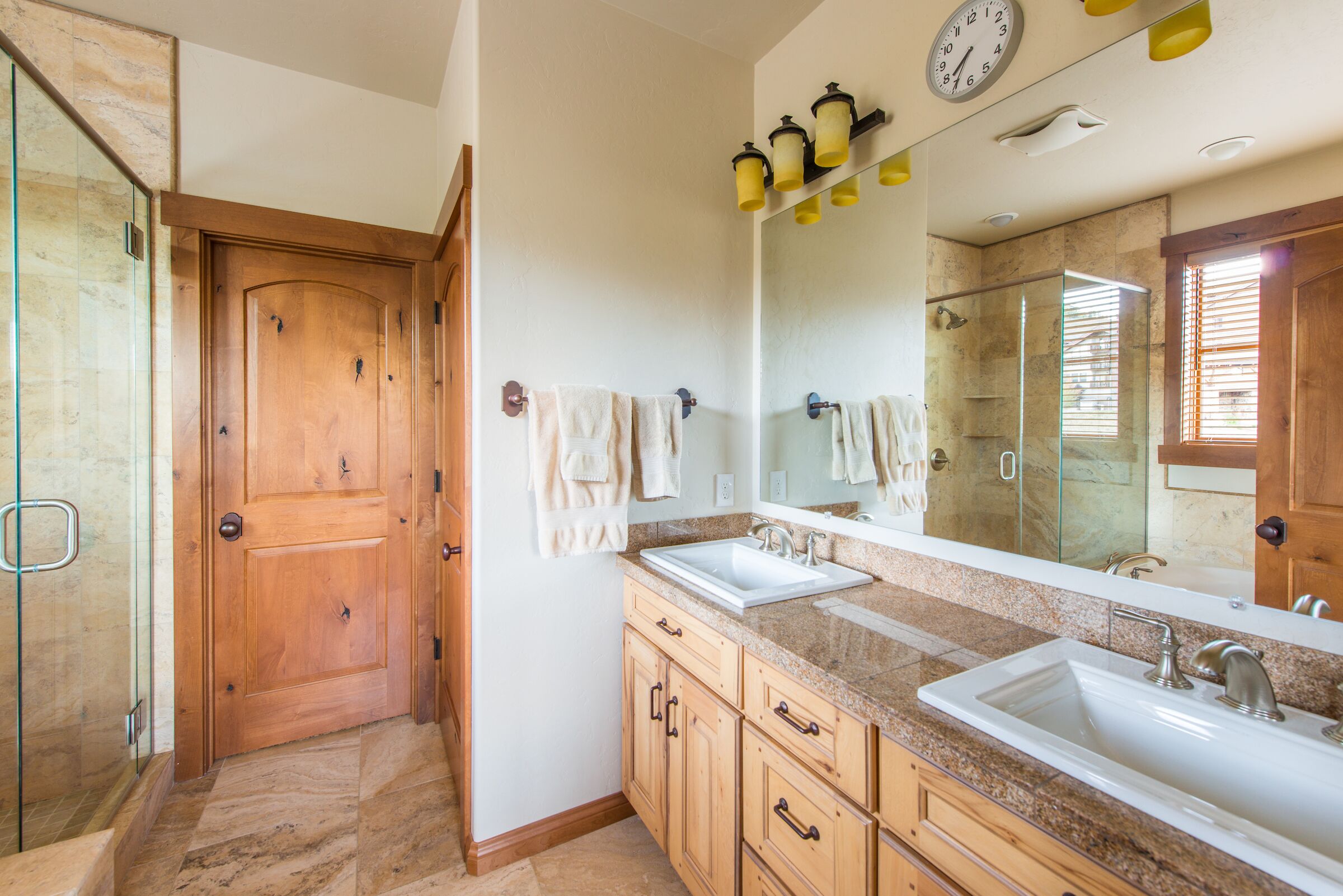 A bathroom with two sinks, wooden cabinets, a glass shower enclosure, and a large mirror with a clock.