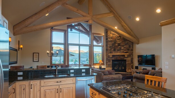 A spacious kitchen with a large island, wooden beams, a stone fireplace, and a view of mountains through large windows. - Hawks Nest Townhome | 3 Bedroom | Mtn Views | Gas Grill | Gas Fireplace (Mt. Crested Butte)