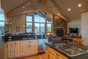A spacious kitchen with a large island, wooden beams, a stone fireplace, and a view of mountains through large windows.