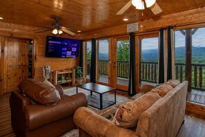 Ein Holzblockhaus-Innenraum mit Wohnzimmer, einem an der Wand montierten Fernseher und einem Balkon mit Bergblick.