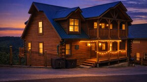 Une cabane en rondins à deux étages, dotée d’un balcon, d’une véranda illuminée de guirlandes lumineuses et d’un toit en métal.