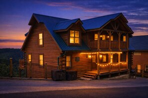 A two-story log cabin with a balcony, a porch with string lights, and a metal roof.