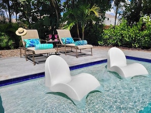 A pool area with two lounge chairs and a table, surrounded by lush greenery. - 667 NEW Heated Pool w Sun Shelf · Blocks2Beach (Naples)