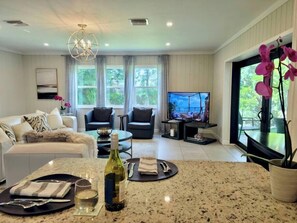 A modern living room with a dining area, a television, and large windows. - 667 NEW Heated Pool w Sun Shelf · Blocks2Beach (Naples)
