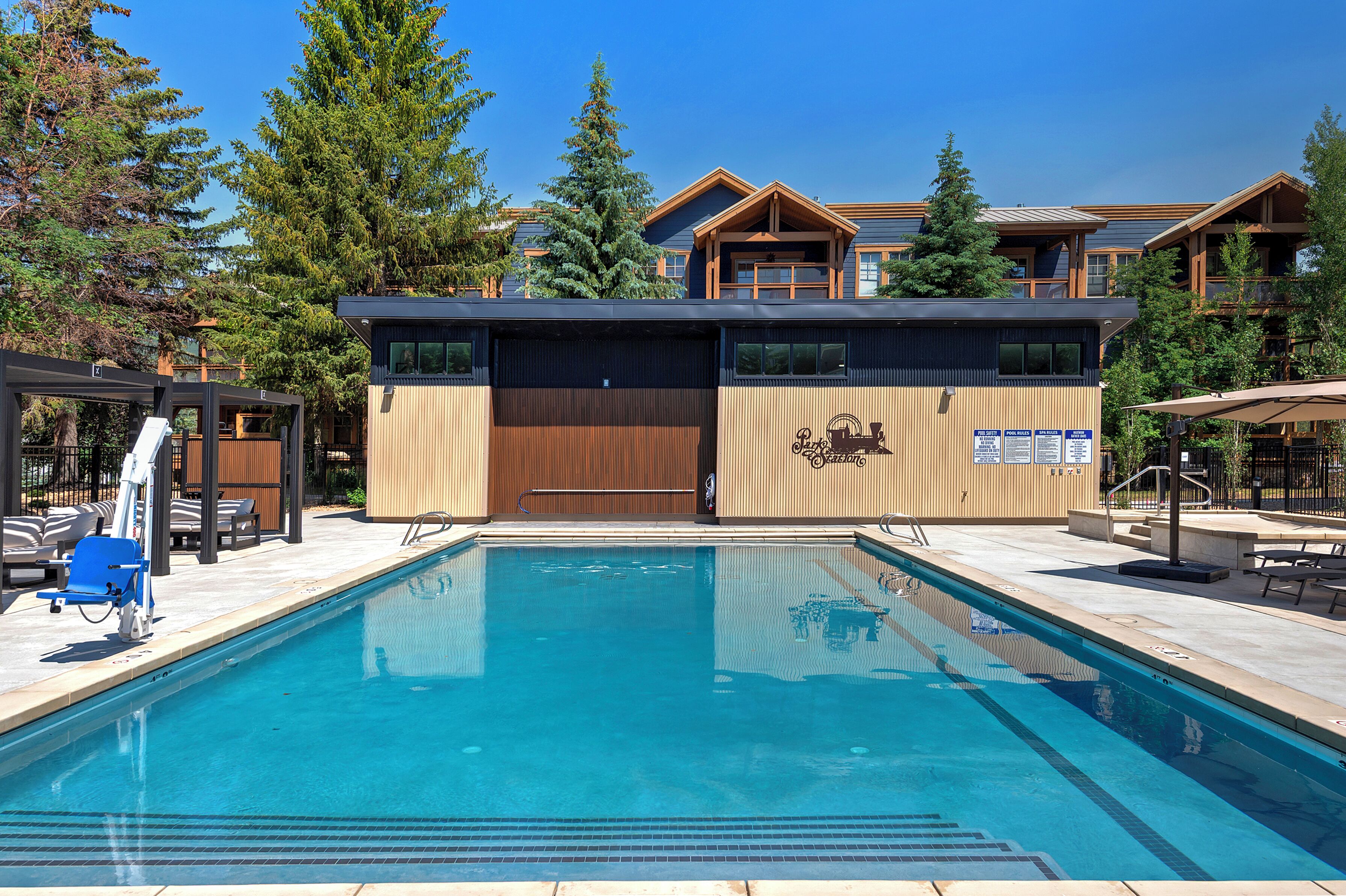 Una piscina con sillas de descanso, un edificio de madera con un telesilla y montañas al fondo.