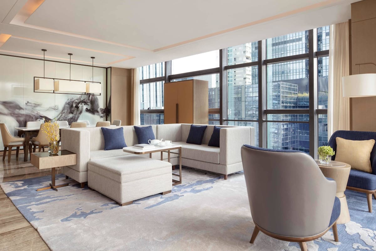 Een moderne woonkamer met een bank, poef en fauteuils, een salontafel en een bijzettafeltje met een vaas. Grote ramen bieden uitzicht op de stad.