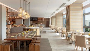 Un restaurante moderno con mesas y bancos de madera, lámparas colgantes y vistas de la ciudad a través de amplios ventanales.