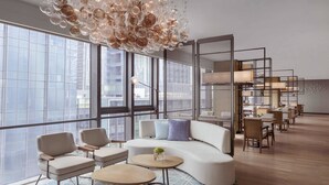 Un área de descanso moderna con un sofá blanco curvo, diversas opciones de asientos y una lámpara colgante original. Amplias ventanas ofrecen vistas de la ciudad.