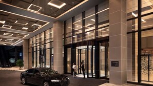 Una entrada de hotel moderna con un coche negro estacionado frente a puertas de vidrio grandes y un letrero que indica el nombre del hotel.