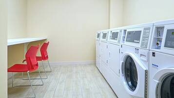 A laundry room with a row of white washing machines and a small table with red chairs.