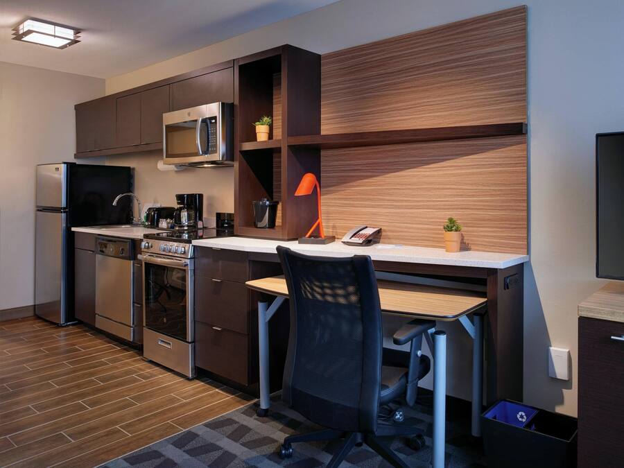 A compact kitchen with a microwave, stove, and refrigerator. There is a wooden desk with a chair and a television.