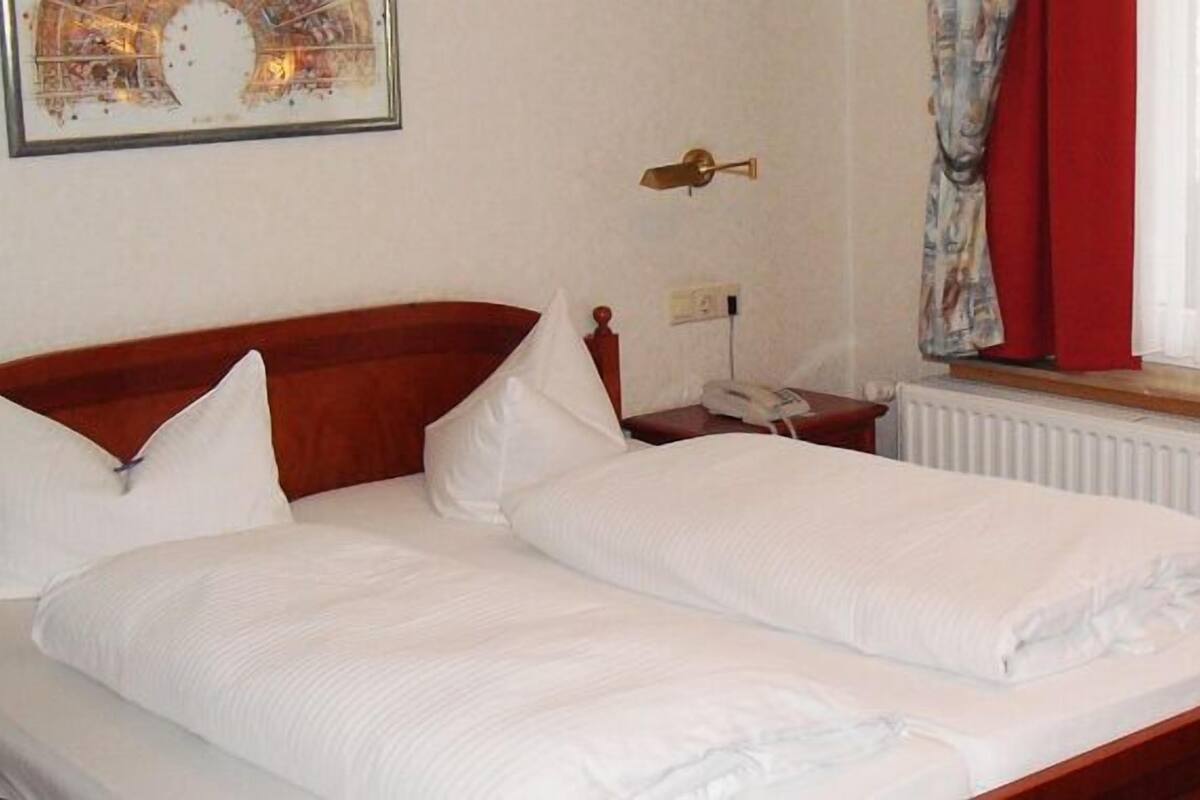 A double bed with white linens in a room with a wooden headboard, a bedside table with a lamp, and a window with sheer curtains.