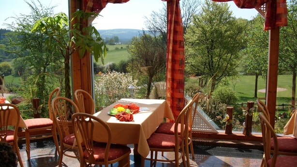 An outdoor dining area with a table set for four, surrounded by wooden chairs with red cushions, overlooking a scenic landscape with trees and a golf course. - Waldhotel (Brilon)