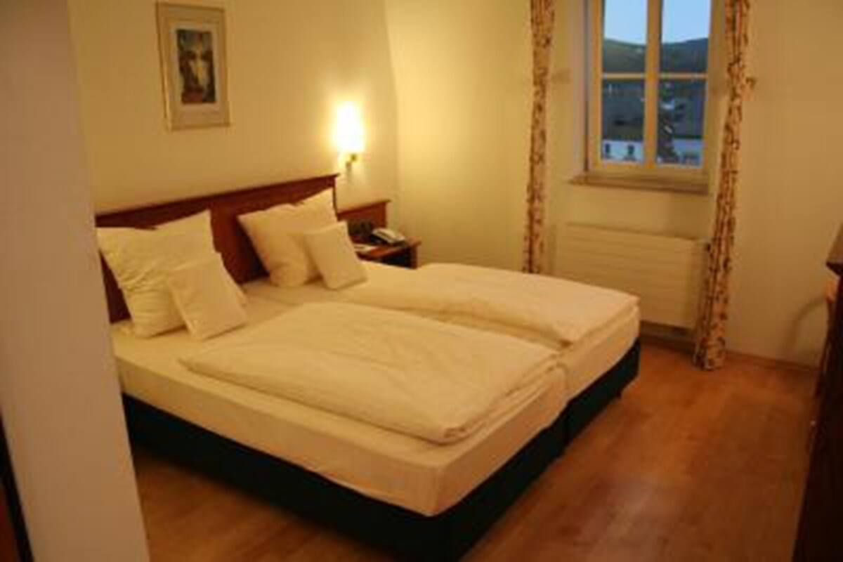 A double bed with white linens, a nightstand with a lamp, and a window with curtains.