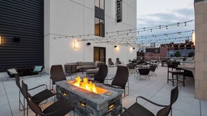 An outdoor patio with a stone fire pit surrounded by chairs, string lights overhead, and a view of the cityscape in the background.