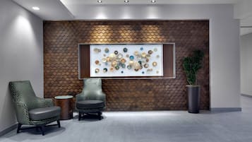 A modern lobby with a geometric patterned wall, a large decorative wall piece, two armchairs, a round table, and a potted plant.