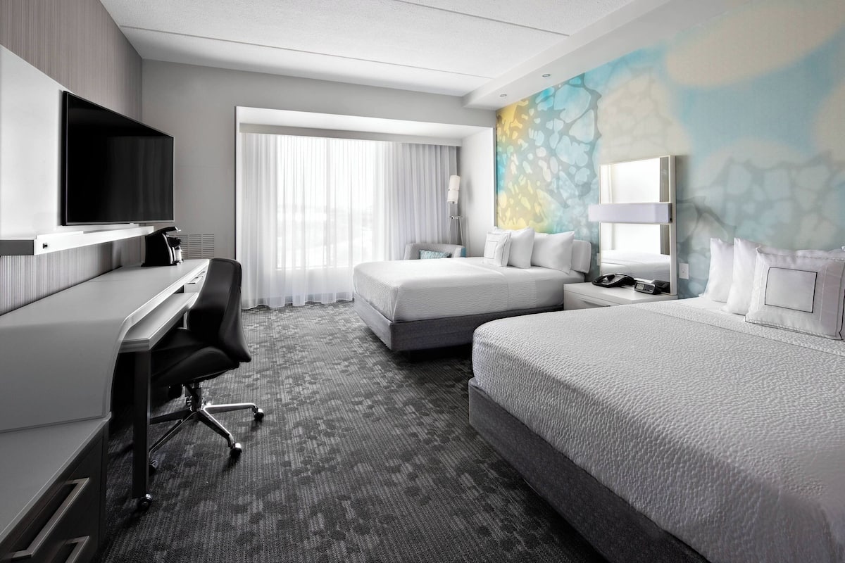 Una camera d'hotel moderna con un letto grande, una scrivania con una sedia, un televisore a schermo piatto e un'ampia finestra con tende leggere.