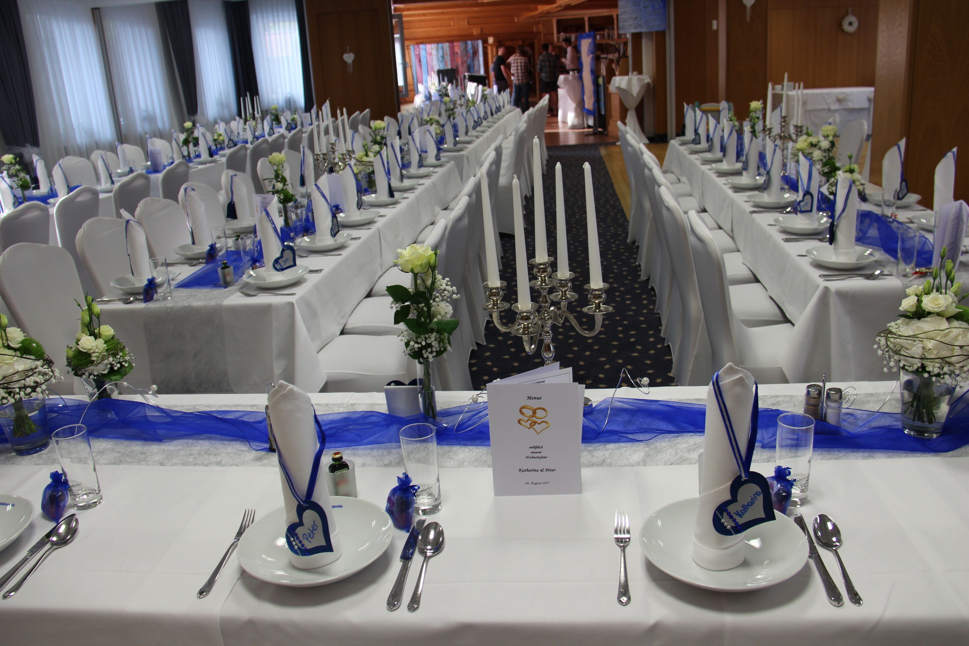 A banquet hall with rows of tables set for a formal event, featuring white tablecloths, blue sashes, and floral centerpieces.