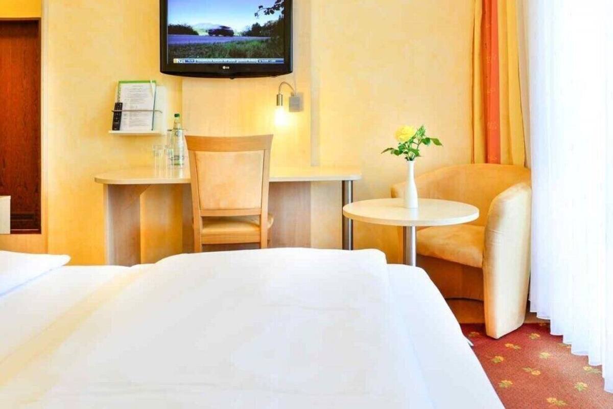 Une chambre d’hôtel comprenant un lit, un bureau, une chaise, une petite table avec un vase, un téléviseur à écran plat fixé au mur et une fenêtre avec des rideaux.