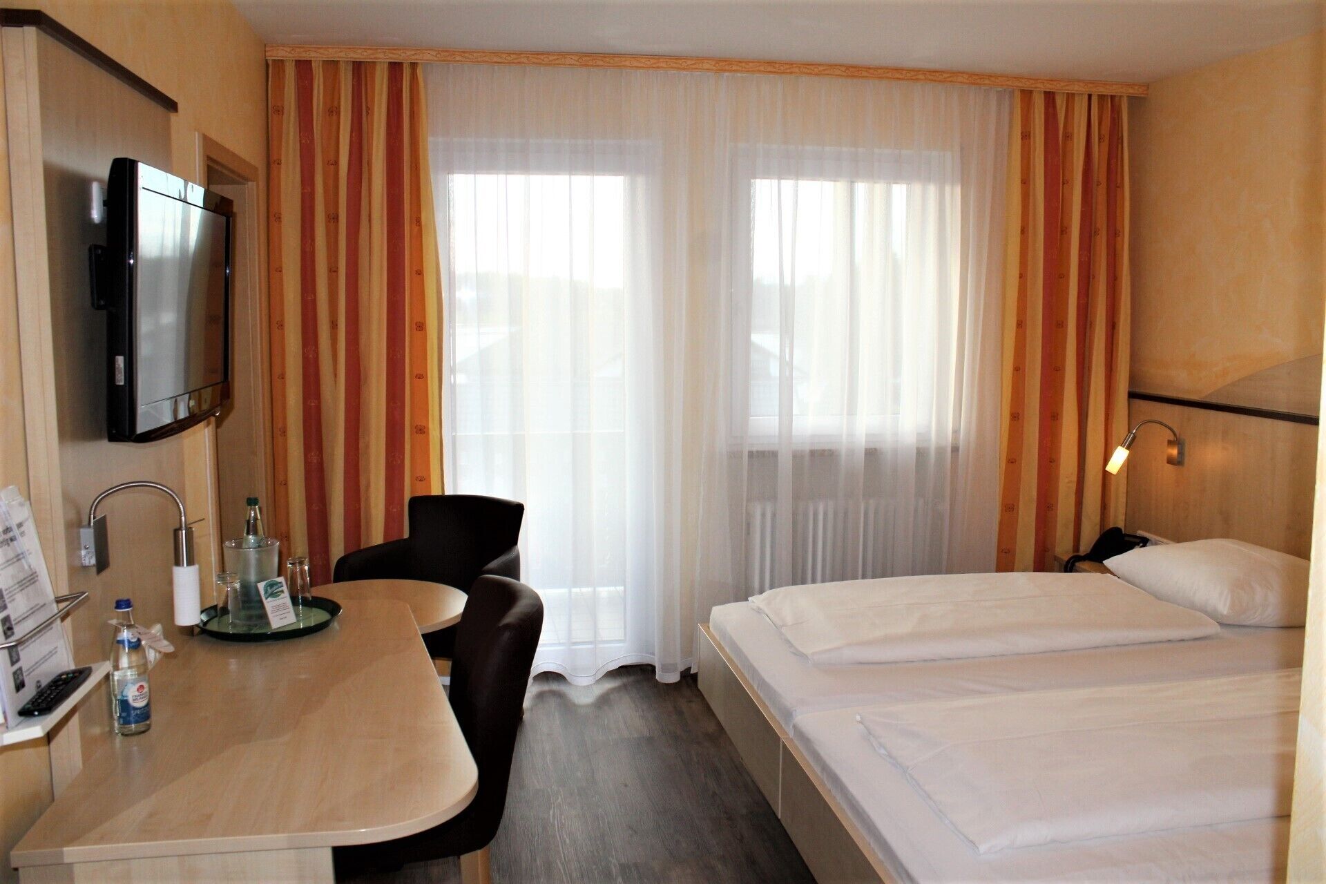 Una habitación de hotel con cama, escritorio con silla, televisor y ventana con cortinas.