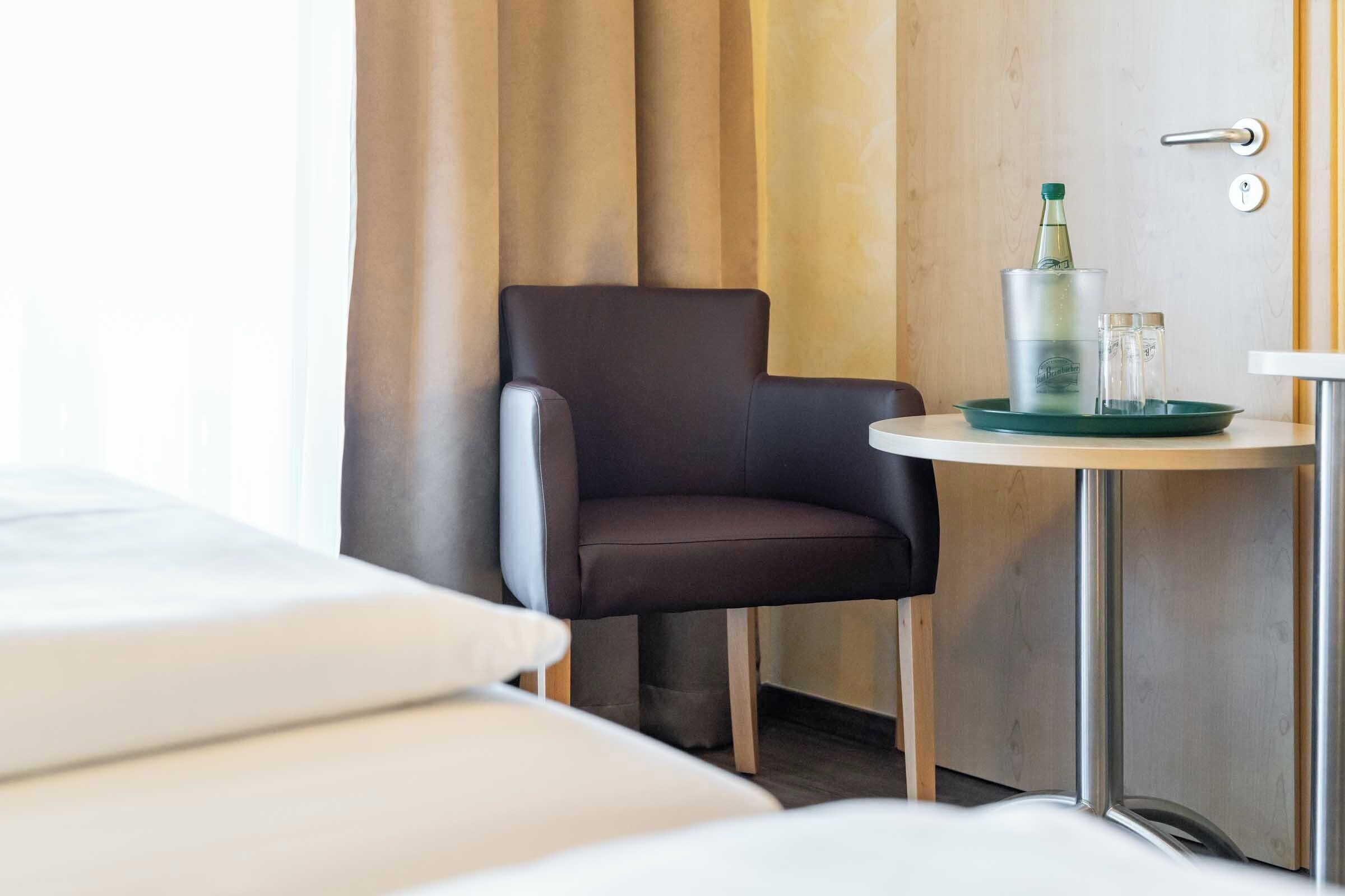 A hotel room with a bed, a chair, a small table with a bottle and glasses, and a door with a handle.