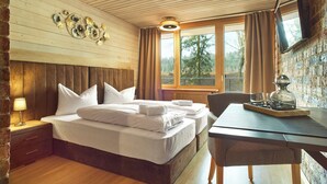 Habitación de hotel con cabecera de madera, cama con ropa blanca, mesita de noche con lámpara, ventana con cortinas y vista a árboles.