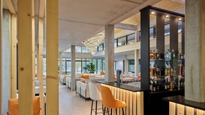 Zona de bar moderna con barra de madera y taburetes, área de estar con sillas blancas y anaranjadas, y un gran ventanal que proporciona luz natural.