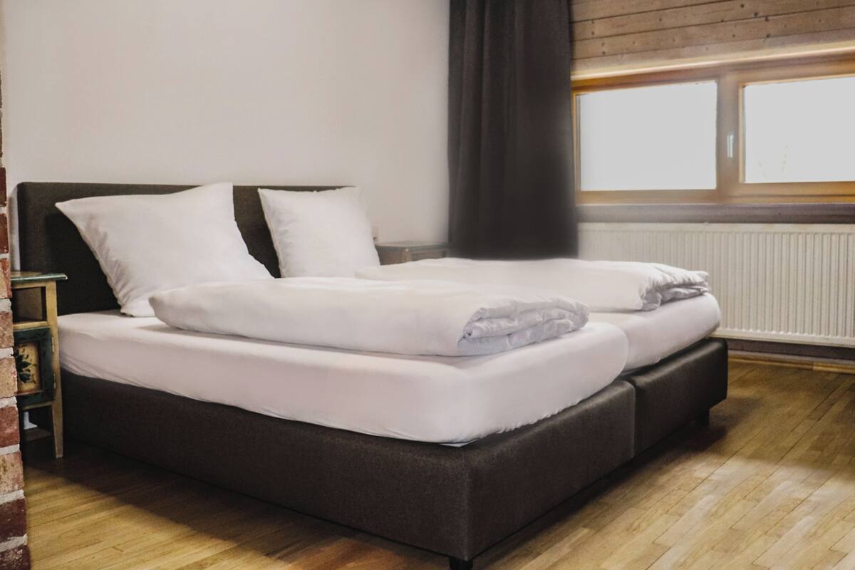 Un llit doble amb roba de llit blanca en una habitació amb terres de fusta i una finestra amb cortines.