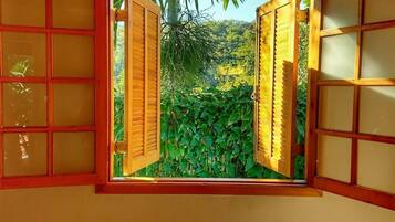 Una habitación con ventana, persianas de madera, vista a áreas verdes y un equipo de aire acondicionado sobre la ventana.
