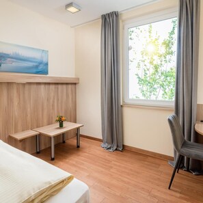 A hotel room with a bed, a desk, a chair, a window with curtains, and a painting on the wall. - Hotel Neumarkt (Berg)