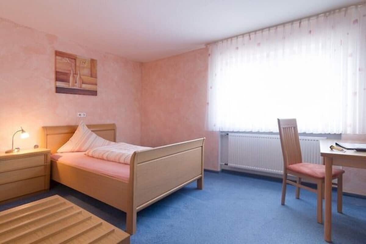 Un dormitorio con cama, mesita de noche, silla y ventana con cortinas.