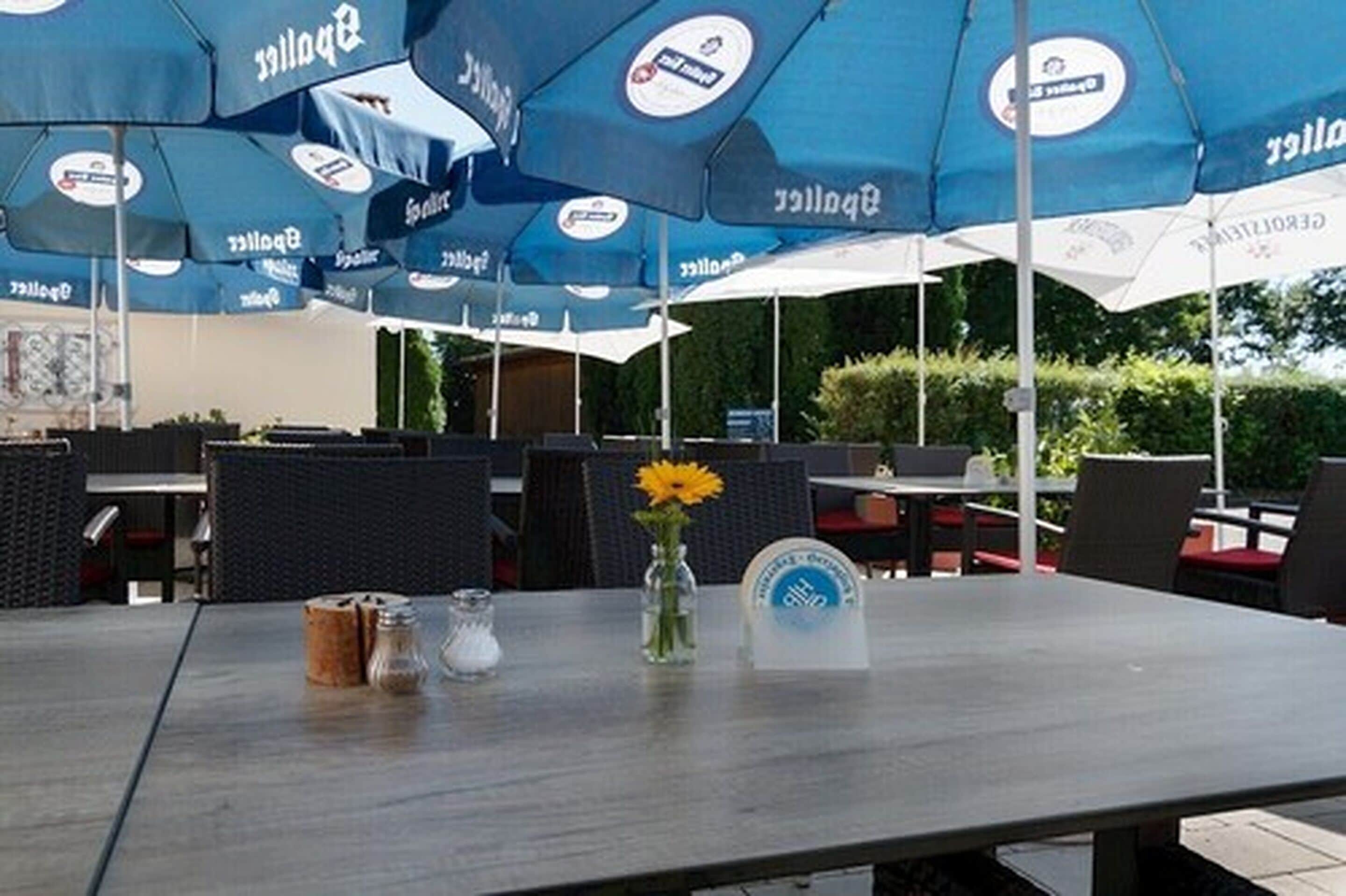 An outdoor seating area with tables and chairs under blue umbrellas, a vase with a yellow flower, and salt and pepper shakers on the table.