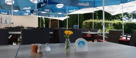 An outdoor seating area with tables and chairs under blue umbrellas, a vase with a yellow flower, and salt and pepper shakers on the table.