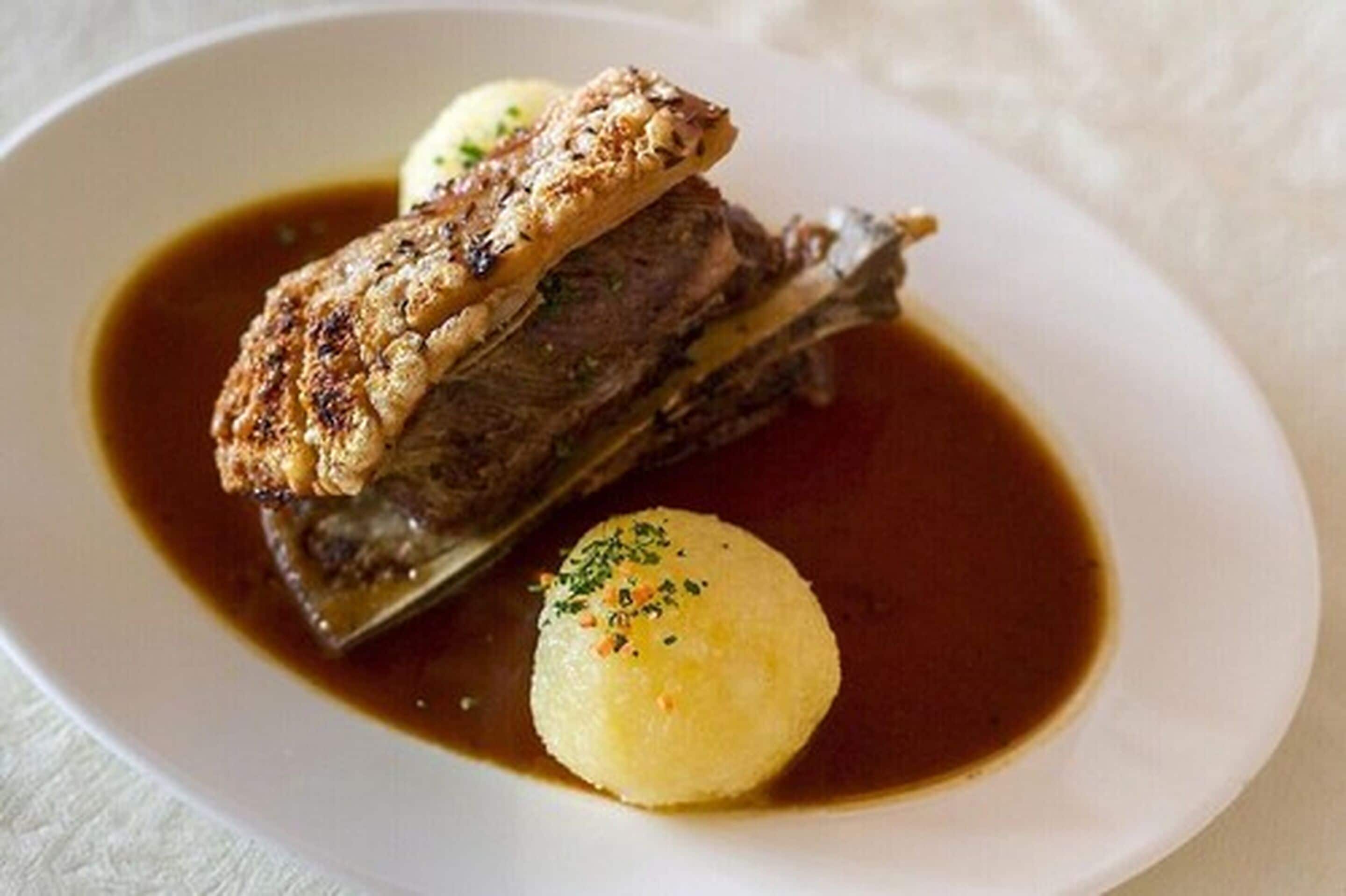 A plate with a piece of meat, possibly beef, topped with a crust, accompanied by two round dumplings and a brown sauce.