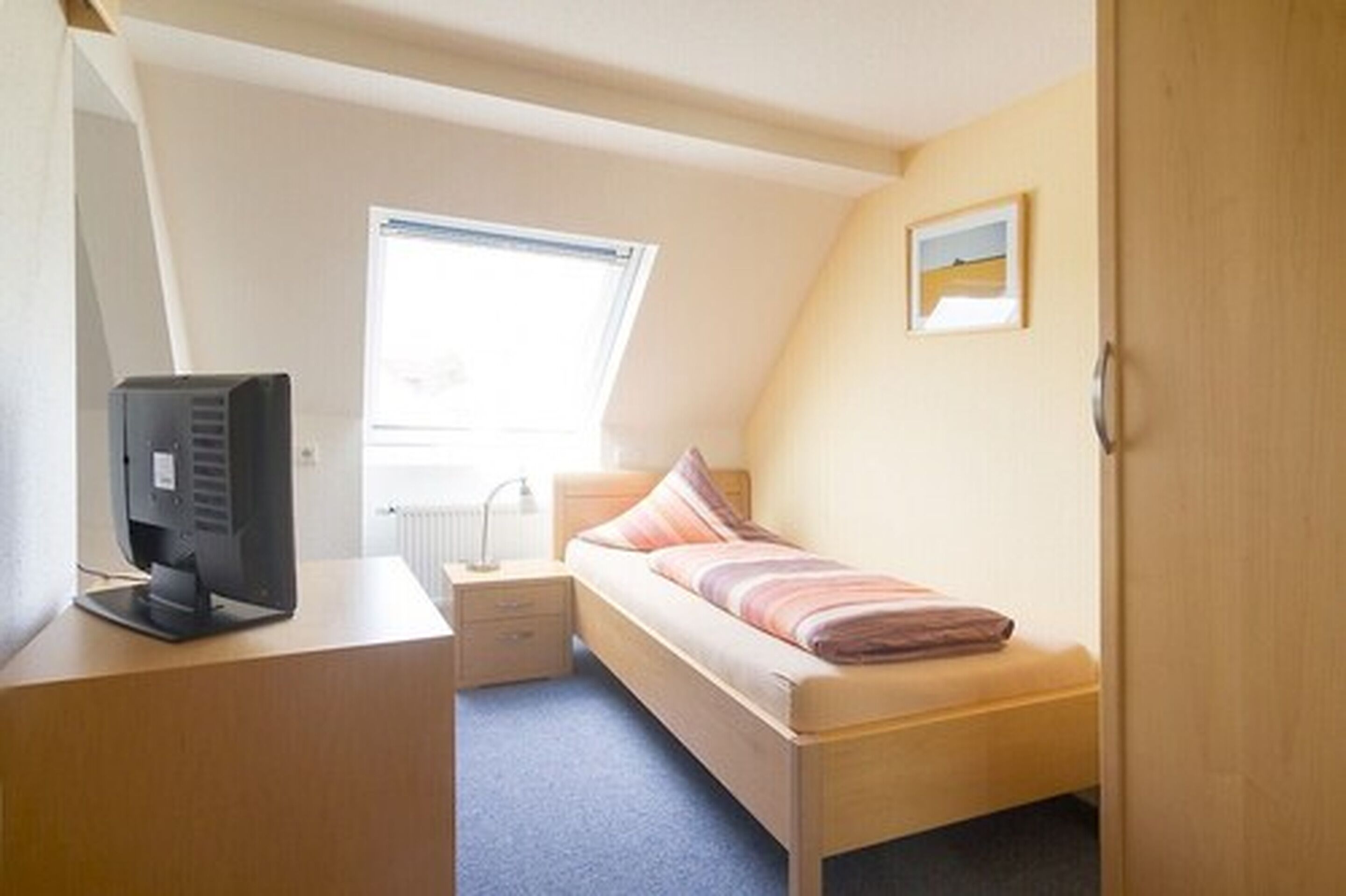 Ein kleiner, gut beleuchteter Raum mit einem Einzelbett, einem Schreibtisch mit einem Computer, einem Fenster, das natürliches Licht hereinlässt, und einem gerahmten Bild an der Wand.