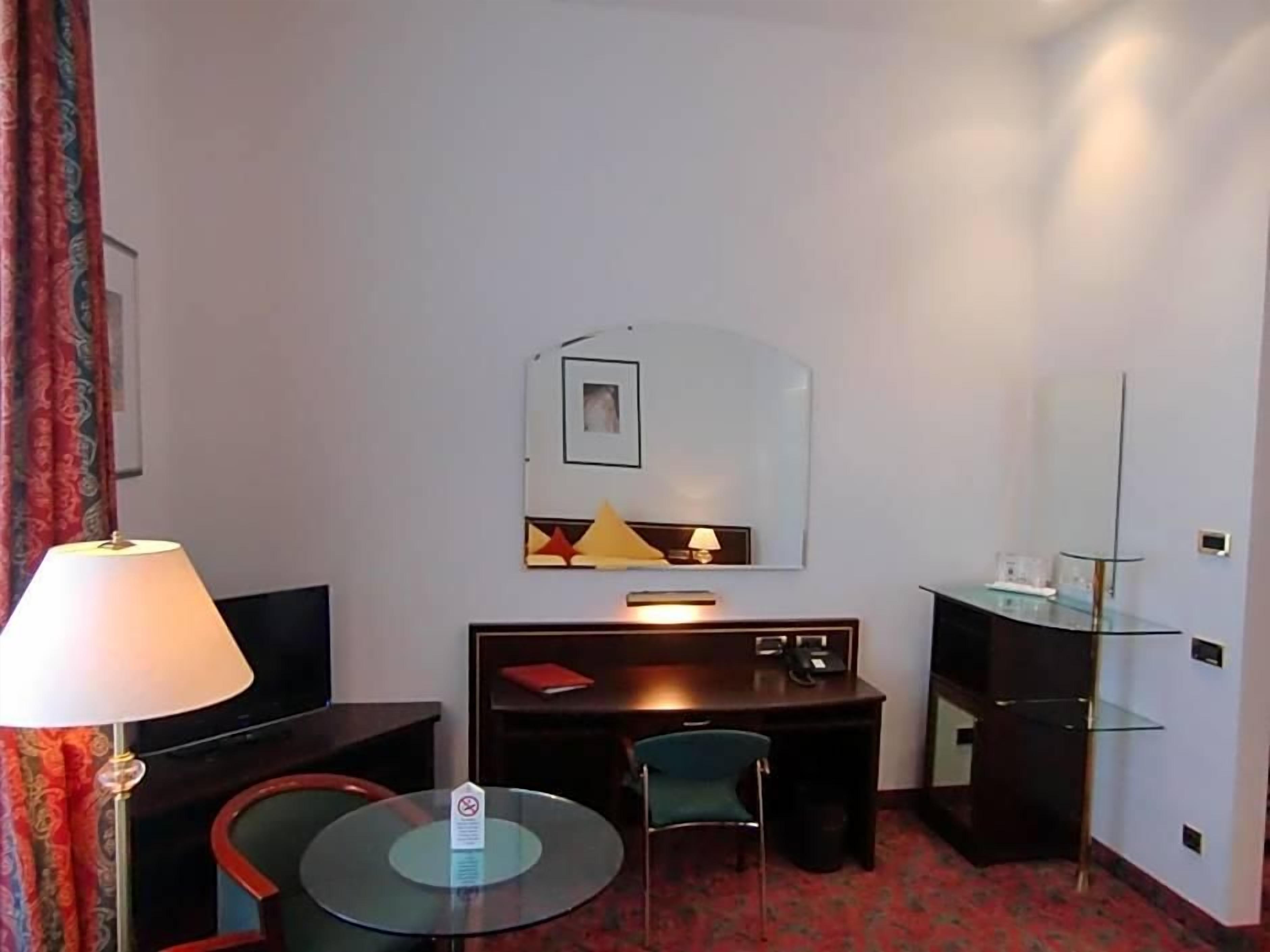 A hotel room with a desk, chair, round table, lamp, and a mirror on the wall.