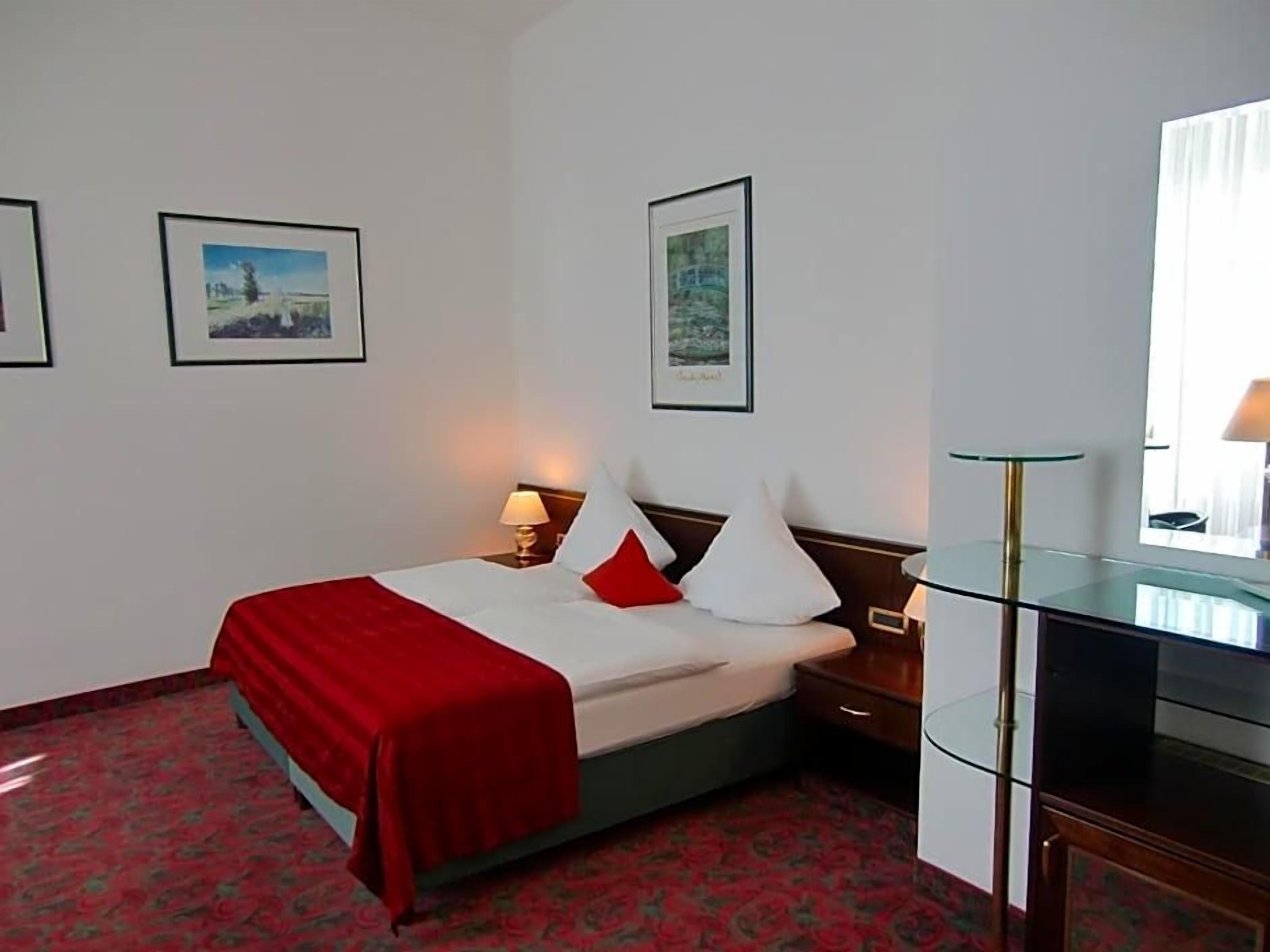 A hotel room with a bed, a desk, and two framed pictures on the wall.