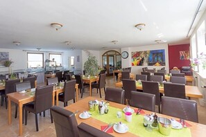 A dining area with multiple tables set for service, a kitchen area in the background, and artwork on the walls.