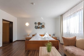 A modern bedroom with a wooden bed, bedside tables, a small table with a plant, and a window with sheer curtains. - Bartholomäus (Zeitlarn)