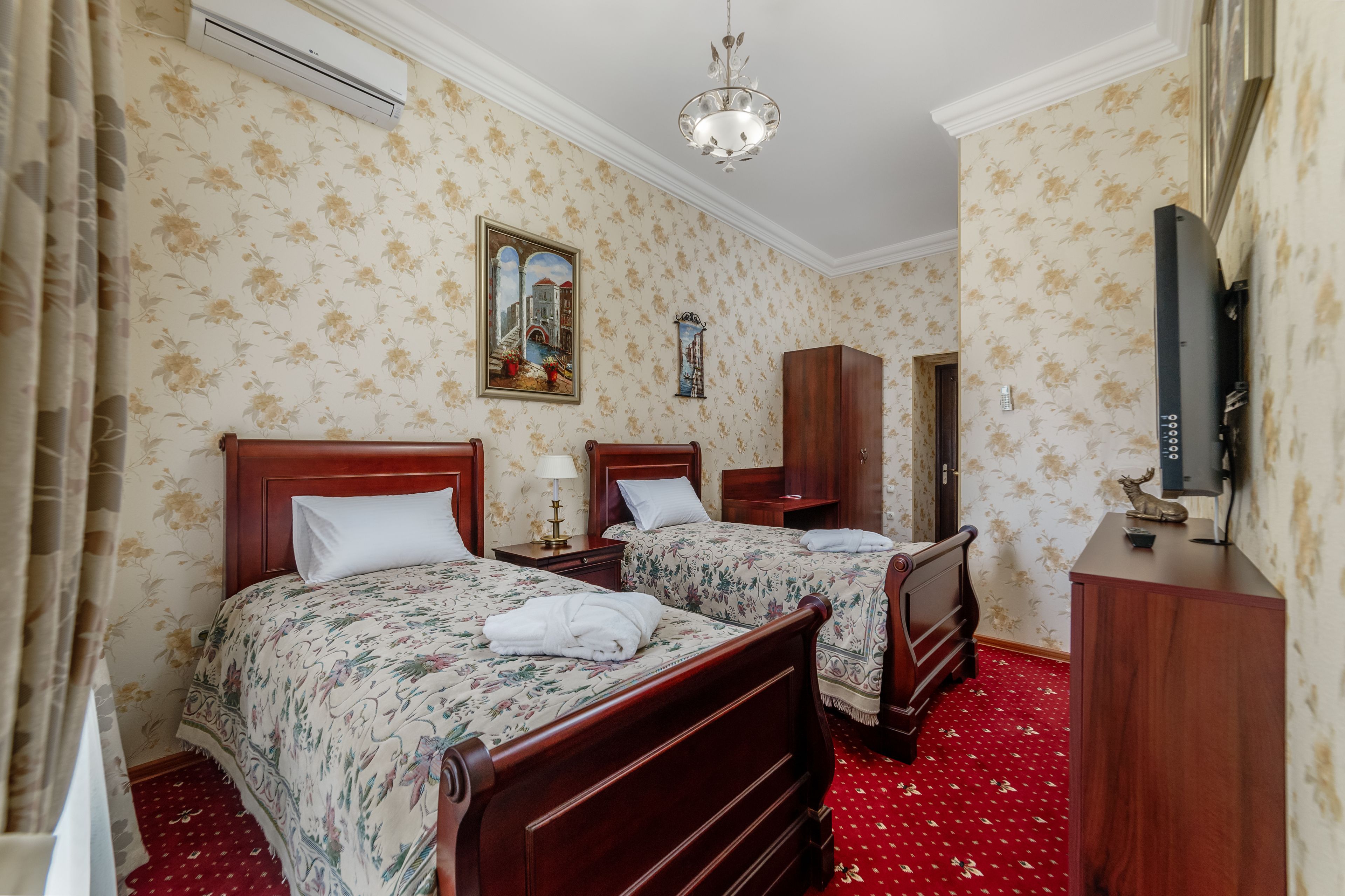 Habitación de hotel con dos camas de madera, alfombra roja, papel tapiz floral, una araña, televisión y un armario.
