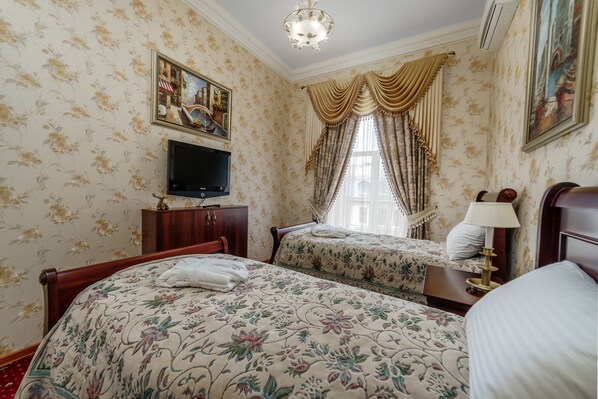 A hotel room with floral wallpaper, two beds, a TV, a chandelier, and a window with curtains.