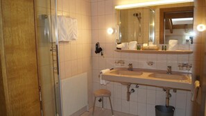 A bathroom with a glass shower enclosure, a double sink, a stool, and a mirror with lights.