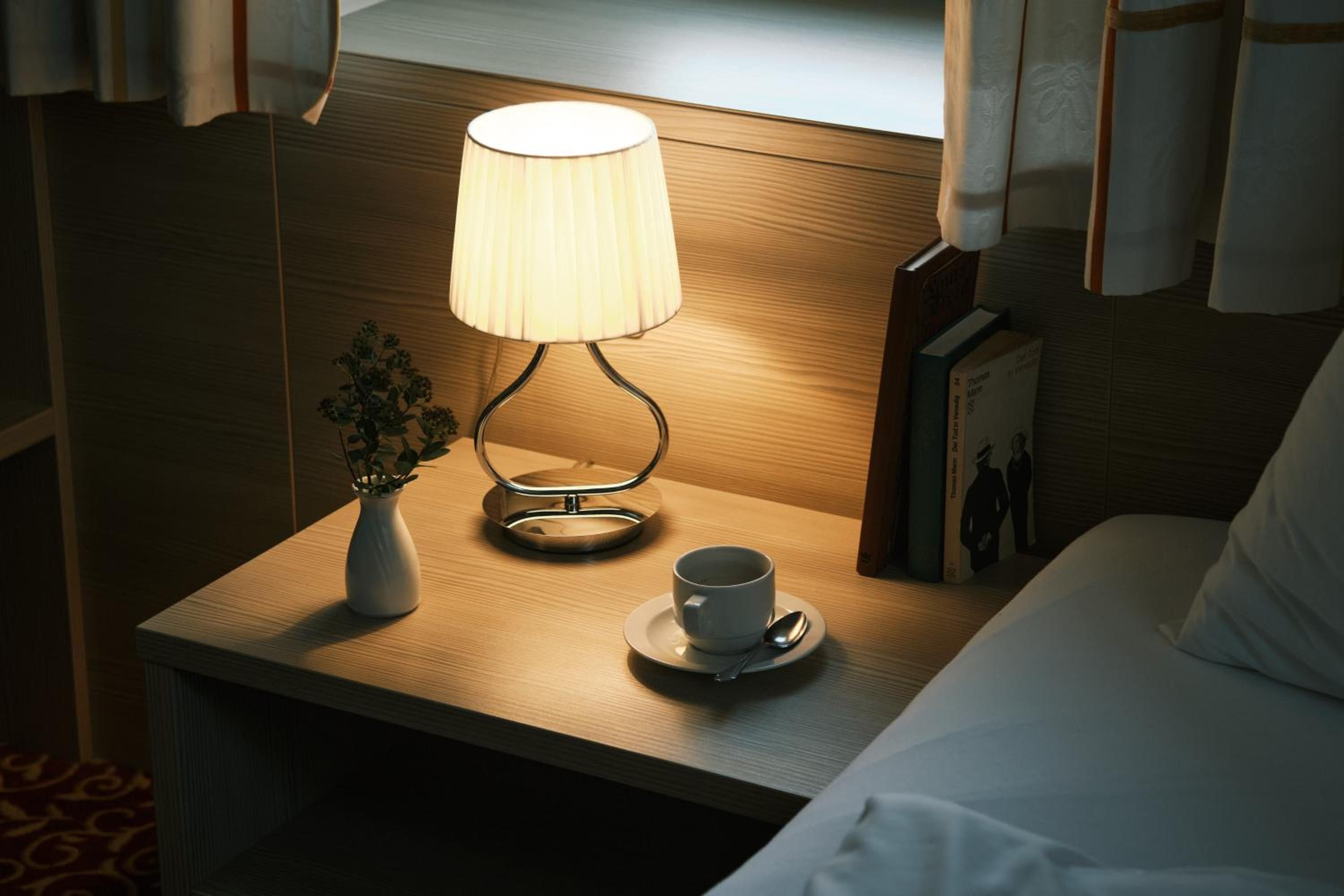 Ein ordentlich bezogenes Bett mit einem weißen Kopfkissen, ein Nachttisch mit einer Lampe, einer Tasse und einer kleinen Pflanze.