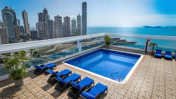 Una piscina en la azotea con sillas reclinables azules, vistas al horizonte urbano y cielo despejado.