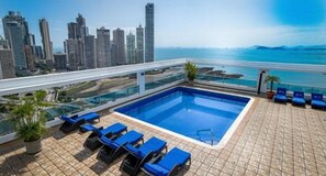 Una piscina en la azotea con sillas reclinables azules, vistas al horizonte urbano y cielo despejado.