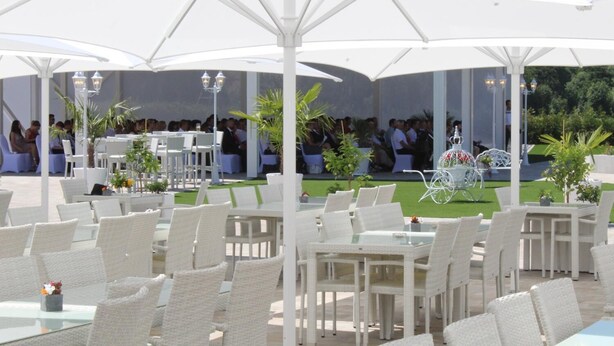 An outdoor dining area with white wicker chairs and tables, covered by large white umbrellas, and surrounded by greenery. - Hotel Grabinger (Schwandorf)